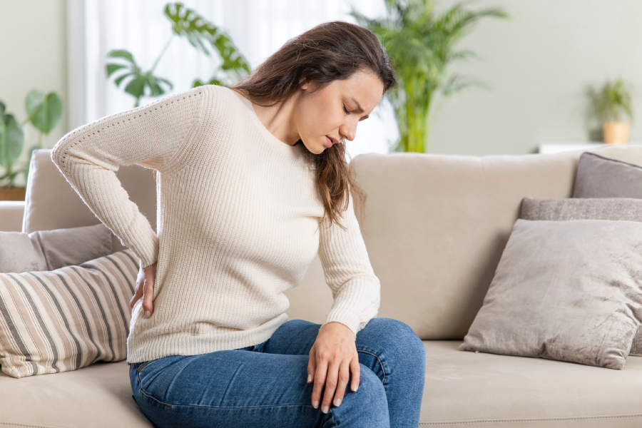 Женщина в светлом свитере сидит на диване и держится за поясницу из-за болей